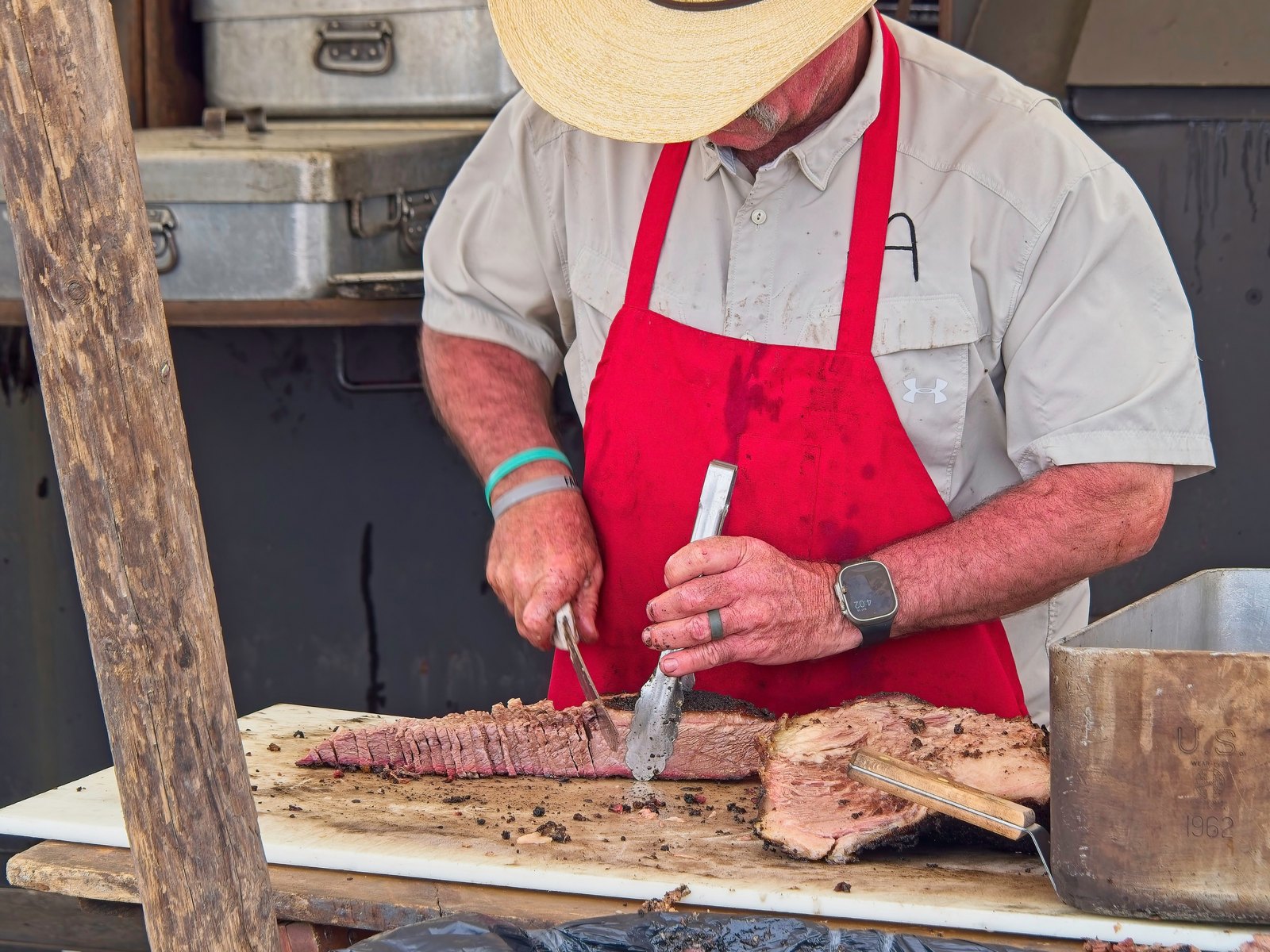 A pitmaster in a straw hat and red apron slicing smoked brisket on a cutting board
