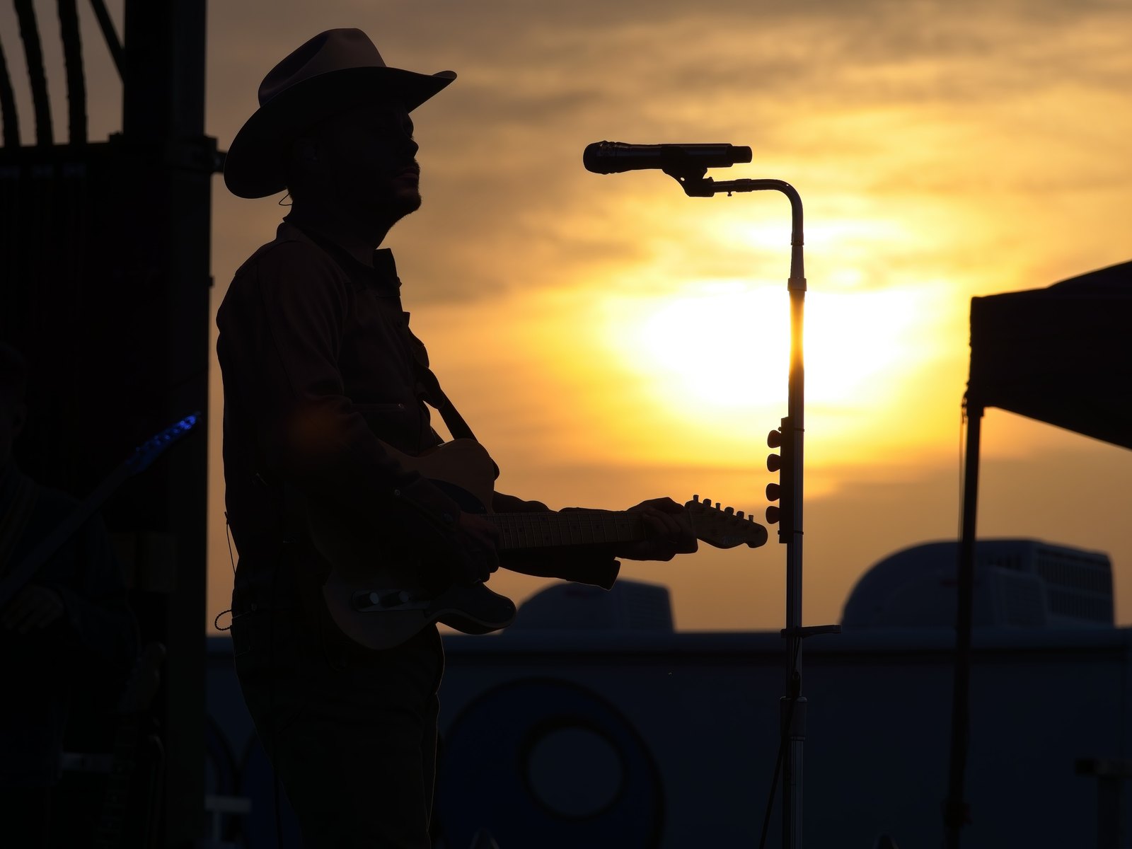 A performer in a cowboy hat silhouetted against a golden sunset behind a microphone stand